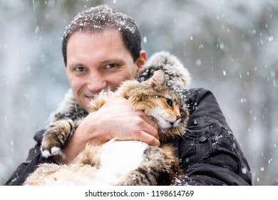 Closeup Of Young Man Holding Scared, Angry Maine Coon Cat Outside, Outdoors In Park In Snow, Snowing Weather During Snowstorm, Storm With Snowflakes, Flakes Falling