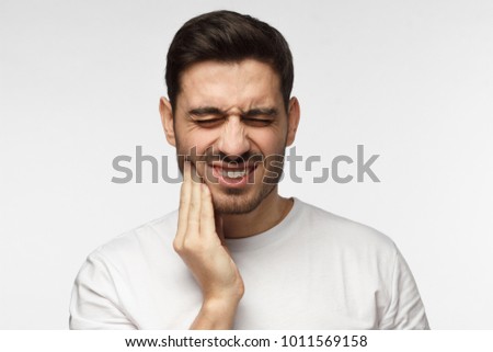 Closeup of young man isolated on gray background touching his face and closing eyes with expression of horrible suffer from health problem and aching tooth, showing dissatisfaction