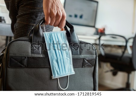 closeup of a young man in an office holding a briefcase and a surgical mask in his hand