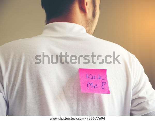 Closeup of a young man with a paper with the text\
kick me attached to his\
back
