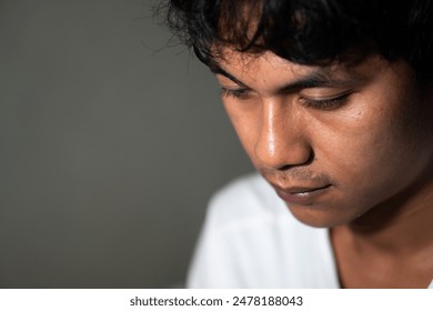 Close-up of a young man with a pensive expression, looking down. He has dark hair and is wearing a white shirt. The background is plain and out of focus. - Powered by Shutterstock