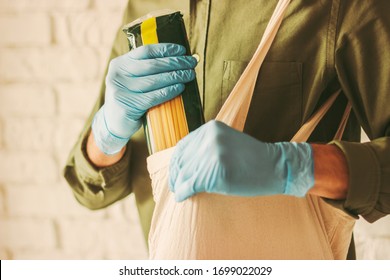 Closeup Of Young Man Shopper In Medical Protective Gloves Putting Macaroni In Cotton Tote Eco Bag In Grocery Store. Hipster Man Shopping In Food Store. Personal Safety, Coronavirus COVID-19 Protection