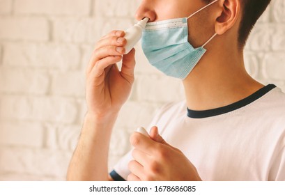 Closeup of young man with sickness using nasal spray for his congested nose. Confident ill doctor in protective medical mask on face using nose drops for runny nose. Coronavirus COVID-19 symptoms