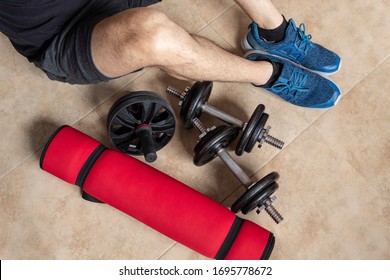 Closeup To A Young Man Sitting On Floor Near To An Iron Dumbbel, An Ab Wheel And A Red Yoga Mat. Elements For Home Workout And Excerside Indoor.  Ideal For Fitness, Sports, And Healthy Life. 