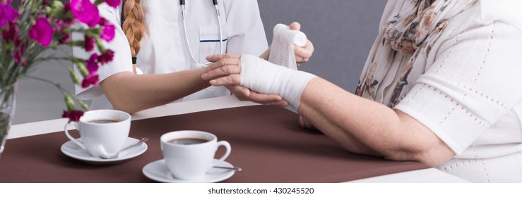 Close-up Of Young Nurse Taking Care Of Older Woman. Carer Dressing A Patient's Wound. Next To Them Two Cups Of Coffee