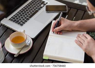 Closeup young programmer sitting at table and taking notes.On table is laptop, phone and cup of tea. Close up of hands.