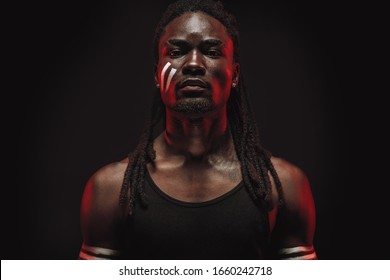 Close-up Of Young Serious Black Male Isolated Over Black Background, Handsome Guy In Black Shirt Stand Seriously Looking At Camera, Confident And Dangerous Person With Indian White Stripes On Cheek