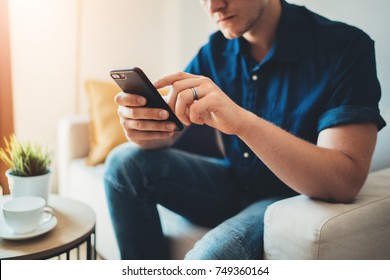 Closeup of young serious man using modern smartphone device while sitting on sofa at home, hipster guy typing an sms message at social network, technology concept