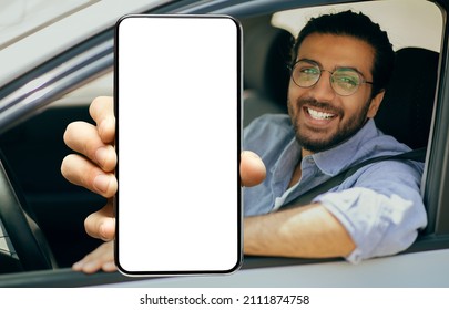 Closeup of young smiling Indian man in glasses showing big mobile phone with white empty screen for mock up sitting on driver seat in new luxury car, holding gadget in hand, selective focus on device
