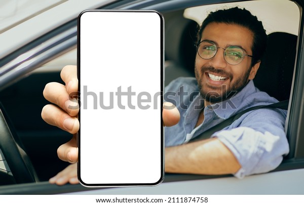 Closeup of young smiling Indian man in glasses\
showing big mobile phone with white empty screen for mock up\
sitting on driver seat in new luxury car, holding gadget in hand,\
selective focus on\
device
