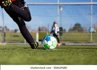 Close-up of young soccer player taking a penalty kick against a young blurred boy acting as goalie in the goal.