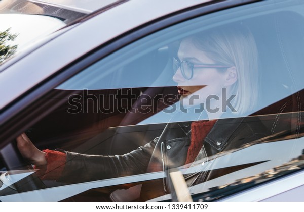 closeup of a young stylish blond woman with\
a red lipstick driving modern car. portrait of female caucasian\
driver. girl learns to drive and\
concentrated