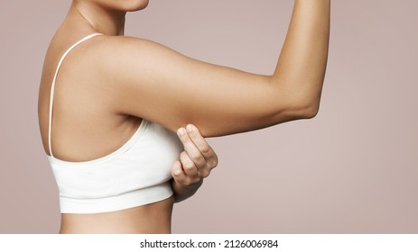 Close-up of a young tanned woman grabbing skin on her upper arm with excess fat isolated on a beige background. Pinching the loose and saggy muscles. Overweight concept