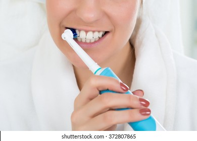 Close-up Of A Young Woman In Bathrobe Brushing Teeth With Electric Toothbrush