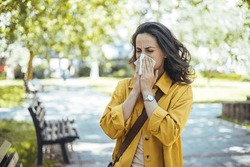 Close-up Of A Young Woman Blowing Nose With Tissue Paper At The Park. Woman With With Allergy Symptom Blowing Nose. Young Pretty Woman Sneezing In Front Of Blooming Tree. Spring Allergy Concept