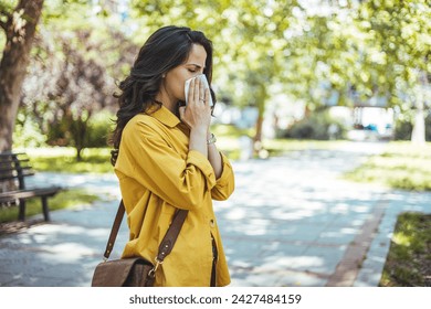 Close-up of a young woman blowing nose with tissue paper at the park. Woman with with allergy symptom blowing nose. Young pretty woman sneezing in front of blooming tree. Spring allergy concept - Powered by Shutterstock