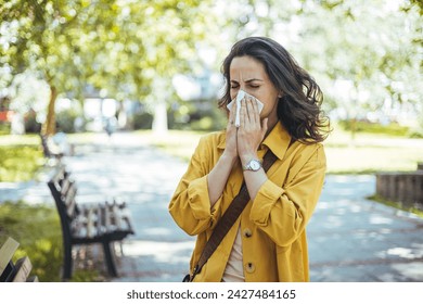 Close-up of a young woman blowing nose with tissue paper at the park. Woman with with allergy symptom blowing nose. Young pretty woman sneezing in front of blooming tree. Spring allergy concept - Powered by Shutterstock