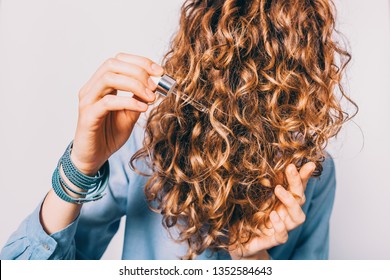 Close-up Young Woman In Blue Shirt Holding Her Curly Brown Hair Applying Moisturizing Oil With Pipette.