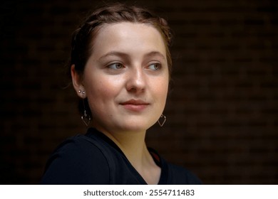 Close-up of a young woman with braided hair and earrings, gazing thoughtfully to the side, set against a textured brick wall, showcasing natural beauty and simplicity. - Powered by Shutterstock