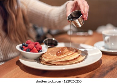 Closeup of young woman at cafe table pouring warm maplesyrup on pancakes