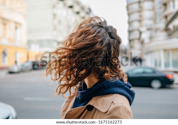 Close-up young woman\
with curly hair wearing beige coat and hoodie standing at city\
street at windy spring\
day.