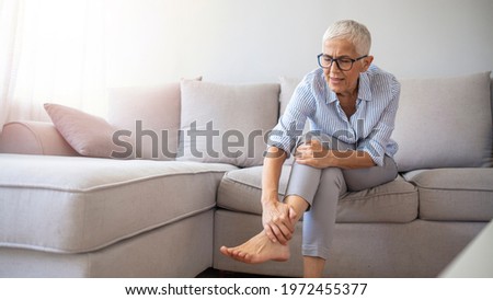  Closeup young woman feeling pain in her foot at home. Healthcare and medical concept. Tired and aching female feet after walking. Woman with feet intense pain sitting on a couch at home.