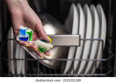 Close-up of young woman hand holding two colored capsule for the dishwasher. In the background, out of focus, is a dishwasher with clean dishes. Small depth of field.