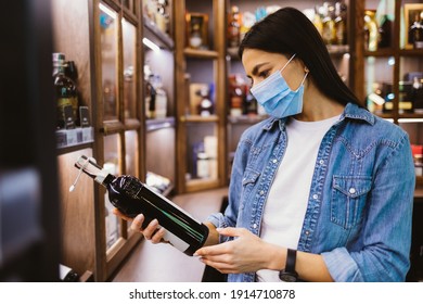 Close-up Young Woman Liquor Store Visitor Wearing Medical Mask For Protection From Virus Holding Bottle Of Wine In Hands, Looking At Label.