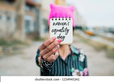 Closeup Of A Young Woman Outdoors Wearing A Pink Pussycat Hat Showing A Notepad In Front Of Her Face With The Text Womens Equality Day Written In It