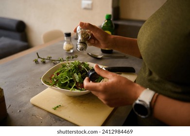 Close-up of a young woman preparing healthy vegetable salad in kitchen, seasoning meal, adding some herbs and salt to bowl, enjoying cooking vegetarian food at home, free space for advertising text - Powered by Shutterstock