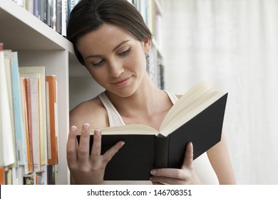 Closeup Of Young Woman Reading By Bookshelves At Home