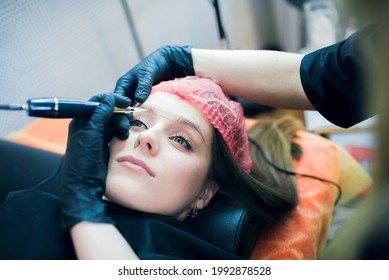 Closeup Of Young Woman's Face With Tatoo Artist's Hands Holding Tatoo Machine Upon Her Brow. Permanent Make Up, Tatoo And Brow Making Professional Sphere.