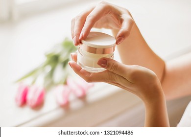 Closeup Of Young Woman's Hand Holding Jar Of Moisturizing Cream In Hands With Spring Flowers Tulips On Background. Gentle Girl Opening Jar With Face Lotion In Arms. Beauty Treatment, Skin Or Body Care