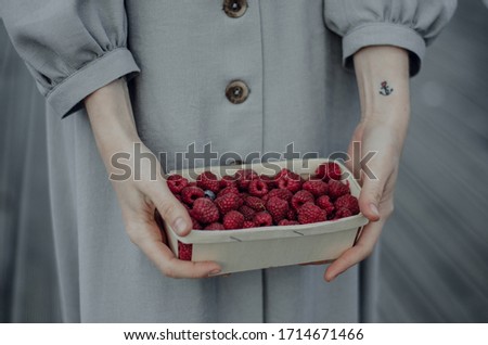 Close-up of young woman's hands holding a wooden basket full of ripe raspberries. Girl in cotton classical dress of gray color with sleeves. Tattooed wrist with anchor. Garden harvest. Soft focus
