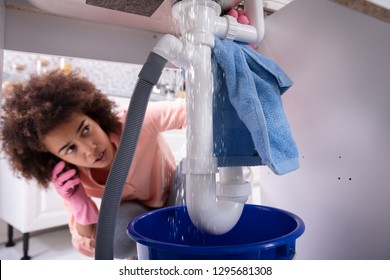 Close-up Of A Young Worried Woman Calling Plumber To Fix Sink Pipe Leakage In Kitchen