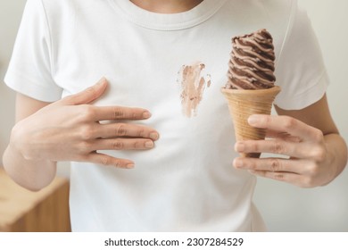 Cloth stain, disappointment asian young woman, girl eating melting ice cream in waffle cone on hot weather, hand show making chocolate cream drop on white t-shirt, spot dirty or smudge on clothes. - Shutterstock ID 2307284529