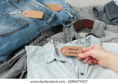 Clothes from females wardrobe on shelf of second hand store. Womans hand holding craft paper tag with inscription second hand. Circular fashion, eco friendly shopping, thrift stores concept