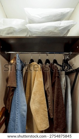clothes hanging on rack inside wardrobe closet with jackets coats and jeans in organized hotel room storage space with pillows on upper shelf warm interior lighting, travel hospitality hotel service