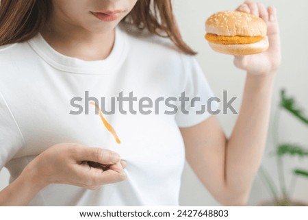 Clothes stain, disappointment asian young woman eat food hand hold hamburger, burger show making chili, tomato sauce smudged spilling, drop on white t-shirt, spot dirty or smudge on clothes, dirt.