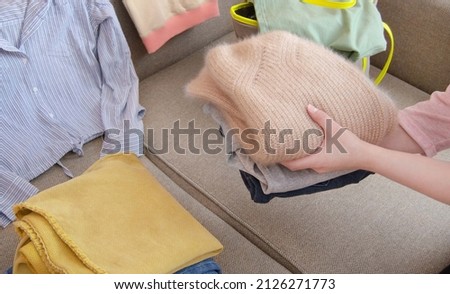 Clothes without a gender attribute are lying on the sofa. A stack of clothes in your hands, preparing clothes at home for donation. Minimalism, donation, consumption of goods, second-hand, spend less.