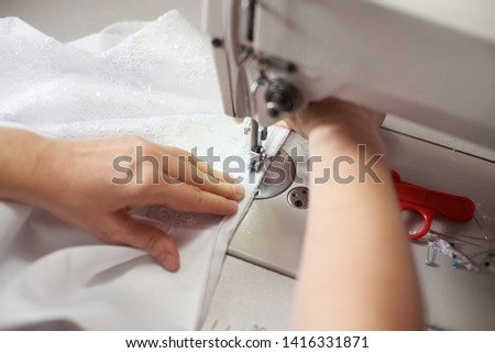 Clothier woman stitching white dress on sewing machine at workplace with pins, threads, scissors on background. Seamstress hands holding and sewing zipper on textile. Blurred background. Close up view