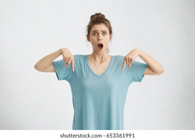 Clothing design and advertisement content. Shocked astonished young Caucasian woman with hair bun looking at camera with mouth fell open, pointing fingers down at her blank blue copyspace t-shirt