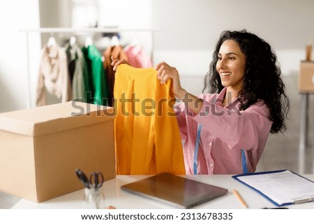 Clothing Design Business. Successful Designer Lady Holding Trendy Garment Packing Order For Buyer Sitting At Table Indoor. Businesswoman Working For Own Online Fashion Store. Ecommerce Concept