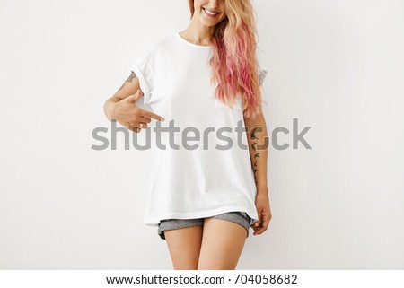 Clothing, design, style, fashion and advertising. Portrait of unrecognizable slim hipster girl with pink hair highlights and tattooed arms smiling, pointing finger at copy space on white black t-shirt