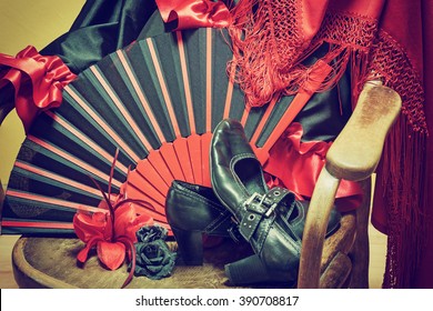 Clothing for Flamenco dance. Black shoes, fan, red scarf with tassels and paper roses are lying on a vintage wooden chair. Edited as a vintage photo with dark edges.