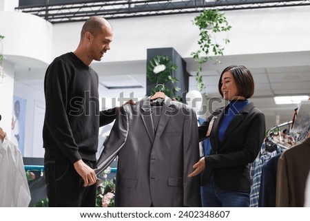 Clothing store asian woman assistant offering advice to an arab man customer, showing formal jacket. Shopping center fashion department consultant and client holding trendy apparel on hanger