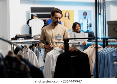 Clothing store employee examining apparel rack and displayed merchandise while using digital tablet. Shopping mall boutique worker doing inventory management with portable gadget - Shutterstock ID 2364449947