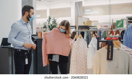 Clothing Store: Female Customer Shopping, Retail Sales Associate Helps. Wearing Protective Face Masks. People In Fashionable Shop, Choosing Stylish Clothes, Colorful Brand With Sustainable Designs