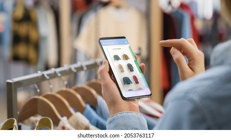 Clothing Store: Female Using Smartphone with Online Fashion Store User Interface to Chek Prices on a Stylish Branded Items. Close Up Over the Shoulder Shot of a Mobile Device. - Shutterstock ID 2053763117