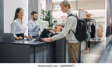 Clothing Store: Handsome Young Man At Checkout Counter Buys Clothes from Friendly Retail Sales Assistant, Paying with Contactless NFC Smartphone Touching Terminal. Shopping Mall with of Designer Brand